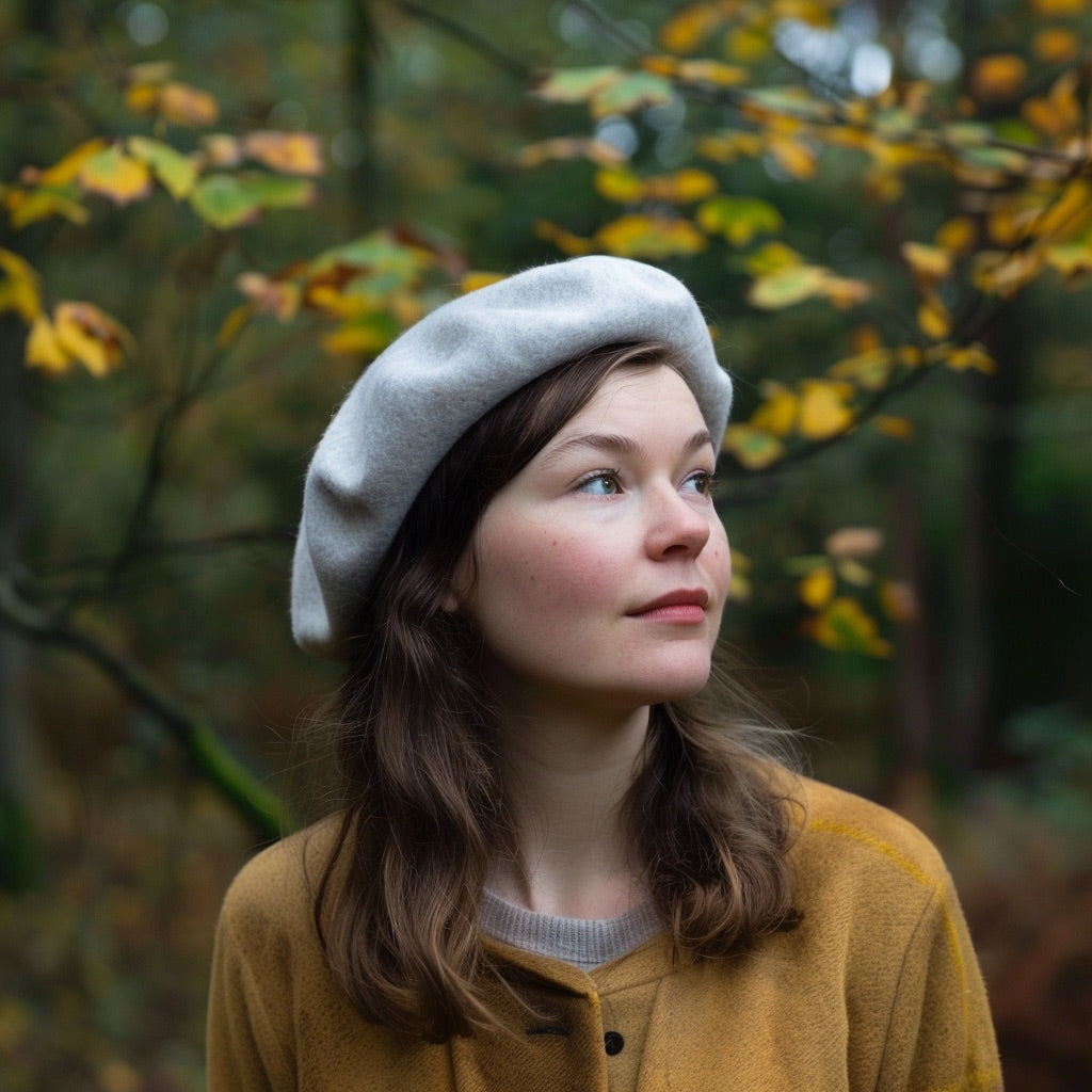 A woman wearing a gray French wool beret, standing in a forest setting with autumn leaves.